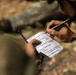 A person in camouflage military uniform is writing on a small notepad in an outdoor, wooded environment. The notepad contains a handwritten military CASEVAC (Casualty Evacuation) 9-Line report format, a standardized medical evacuation request used in military operations. The person is using a Staedtler Lumocolor permanent marker/pen to fill in the form.