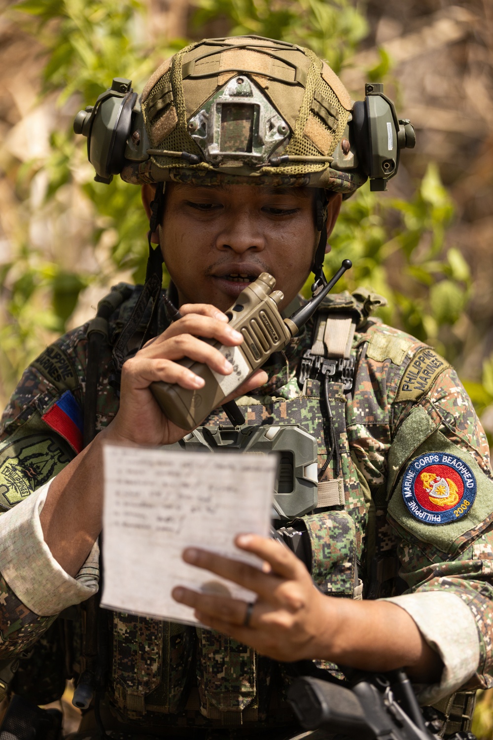 A Philippine Marine soldier is captured in the field reading from a handwritten notepad while communicating via a handheld tactical radio. The soldier is wearing full combat gear including a FAST helmet with ear protection and NVG mount, digital camouflage uniform, and tactical vest. Patches on the uniform identify the individual as a member of the Philippine Marines and the Marine Corps Beachhead unit (2008).