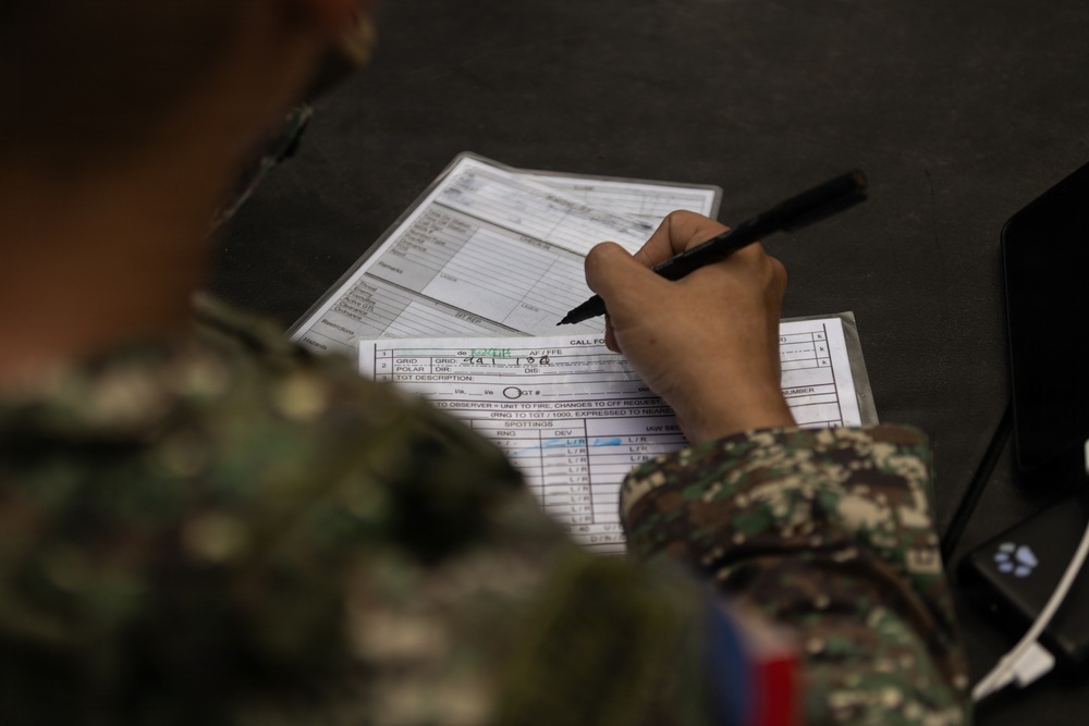 A military personnel in camouflage uniform is writing on official military forms placed on a clipboard or flat surface. The forms appear to be Call For Fire (CFF) request documents, which are standard artillery fire mission coordination sheets. The soldier is actively filling in data fields using a black marker or pen.