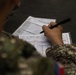 A military personnel in camouflage uniform is writing on official military forms placed on a clipboard or flat surface. The forms appear to be Call For Fire (CFF) request documents, which are standard artillery fire mission coordination sheets. The soldier is actively filling in data fields using a black marker or pen.