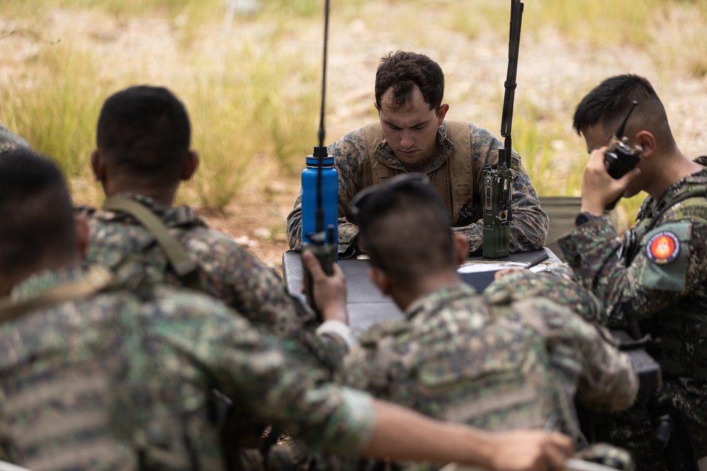A group of military personnel from at least two different nations are gathered around a field table in an outdoor environment, appearing to conduct a joint planning or coordination session. A central figure in US Marine Corps MARPAT camouflage reviews documents while others hold radios and reference materials. The setting and mixed uniforms suggest a multinational military exercise or training operation.