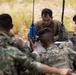 A group of military personnel from at least two different nations are gathered around a field table in an outdoor environment, appearing to conduct a joint planning or coordination session. A central figure in US Marine Corps MARPAT camouflage reviews documents while others hold radios and reference materials. The setting and mixed uniforms suggest a multinational military exercise or training operation.