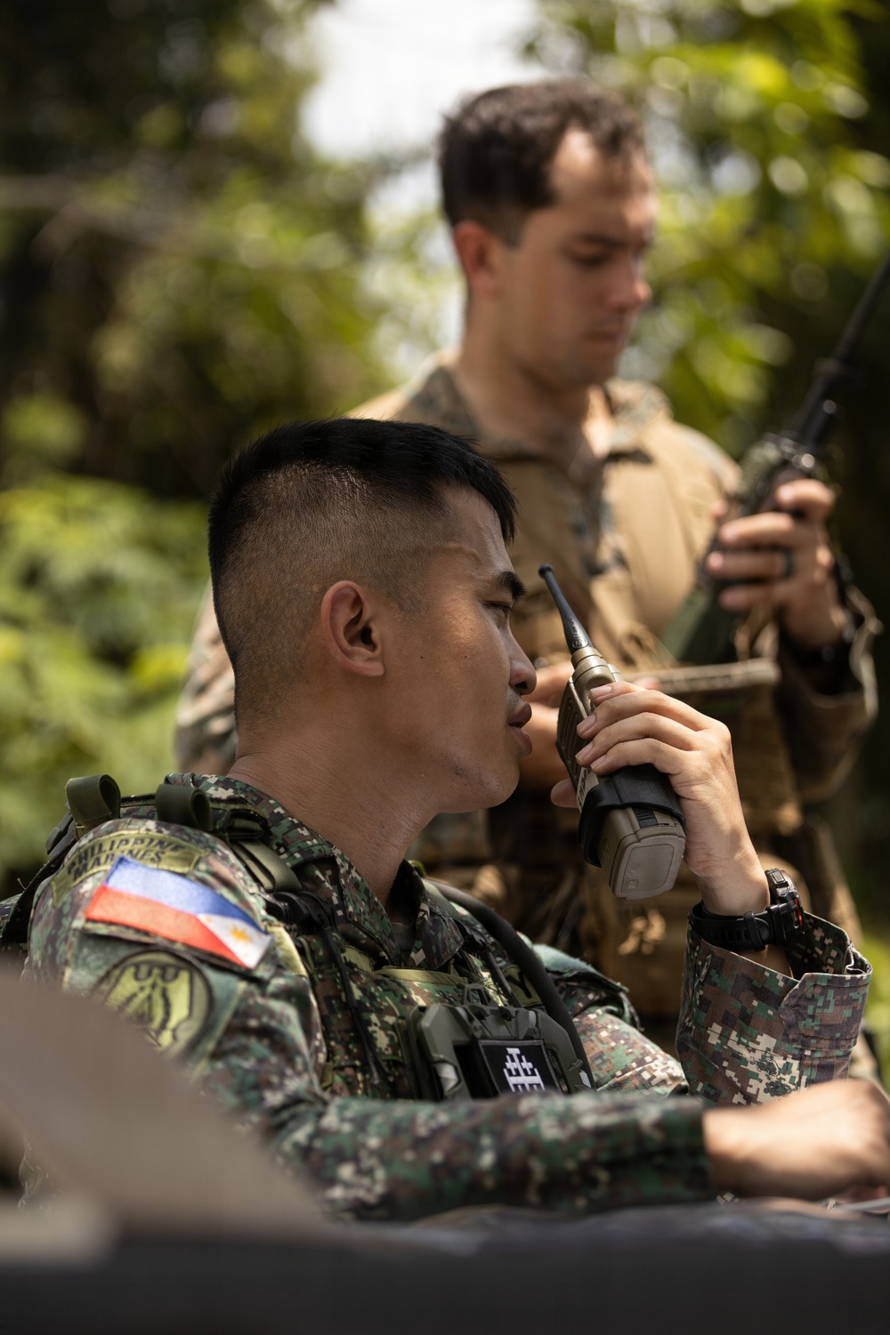 Two military personnel are engaged in radio communications in an outdoor, vegetated setting. The foreground soldier wears Philippine Armed Forces digital camouflage with a visible Philippine flag patch and is speaking into a handheld radio. A second soldier in the background, wearing what appears to be US Marine Corps MARPAT camouflage, holds a separate radio device.