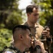 Two military personnel are engaged in radio communications in an outdoor, vegetated setting. The foreground soldier wears Philippine Armed Forces digital camouflage with a visible Philippine flag patch and is speaking into a handheld radio. A second soldier in the background, wearing what appears to be US Marine Corps MARPAT camouflage, holds a separate radio device.