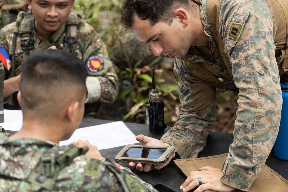 Military personnel from what appears to be two different nations are gathered around a field table in an outdoor jungle or forested environment. A soldier in MARPAT (US Marine) camouflage is leaning over and pointing at a ruggedized tablet device held by another soldier. The scene suggests a joint military training exercise or operational briefing involving digital equipment.