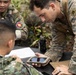 Military personnel from what appears to be two different nations are gathered around a field table in an outdoor jungle or forested environment. A soldier in MARPAT (US Marine) camouflage is leaning over and pointing at a ruggedized tablet device held by another soldier. The scene suggests a joint military training exercise or operational briefing involving digital equipment.