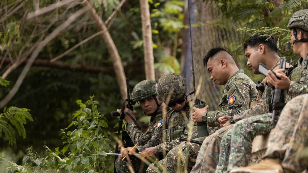 A group of military personnel in camouflage uniforms are gathered in a wooded outdoor setting, appearing to conduct a briefing or planning session. Several soldiers are seated and reviewing documents or notes, while at least one holds a radio communication device. The scene suggests a field exercise or operational planning activity.