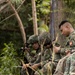 A group of military personnel in camouflage uniforms are gathered in a wooded outdoor setting, appearing to conduct a briefing or planning session. Several soldiers are seated and reviewing documents or notes, while at least one holds a radio communication device. The scene suggests a field exercise or operational planning activity.