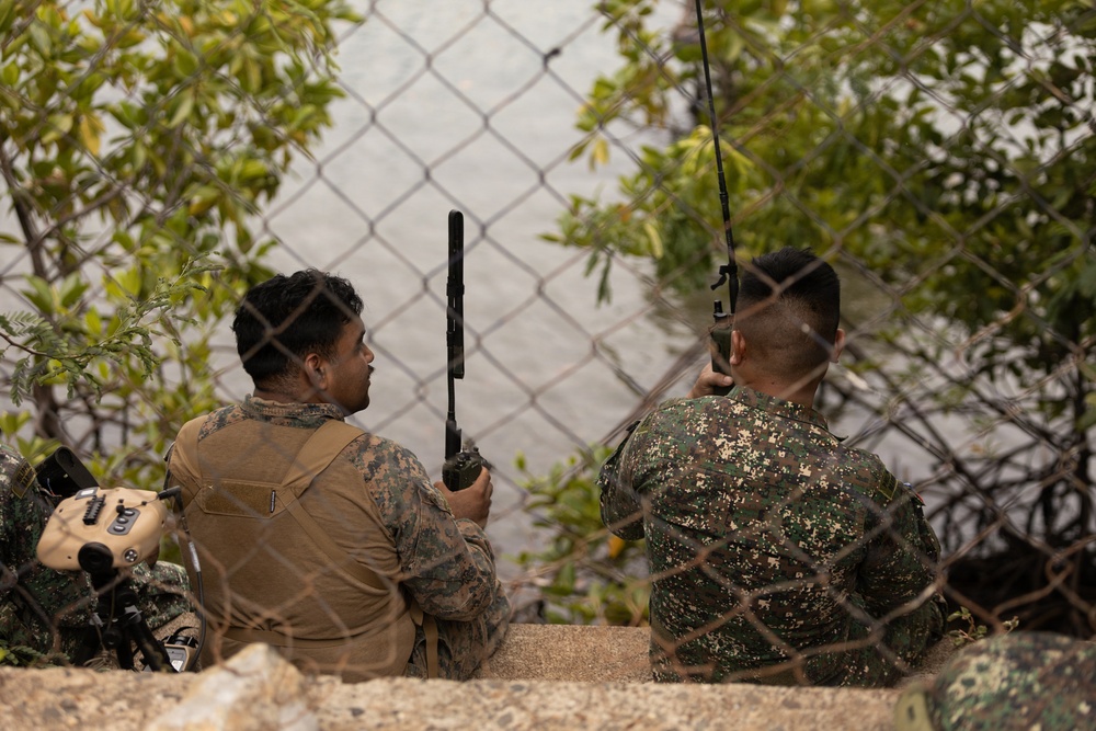 Two military personnel in different camouflage uniforms are seated behind a chain-link fence near a waterway, both holding radio communication devices with extended antennas. The soldier on the left wears MARPAT (US Marine Corps pattern) camouflage with a tan plate carrier vest, while the soldier on the right wears a green digital camouflage pattern suggesting a different military branch or nation. They appear to be conducting a coordinated communications or observation exercise.