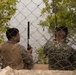 Two military personnel in different camouflage uniforms are seated behind a chain-link fence near a waterway, both holding radio communication devices with extended antennas. The soldier on the left wears MARPAT (US Marine Corps pattern) camouflage with a tan plate carrier vest, while the soldier on the right wears a green digital camouflage pattern suggesting a different military branch or nation. They appear to be conducting a coordinated communications or observation exercise.