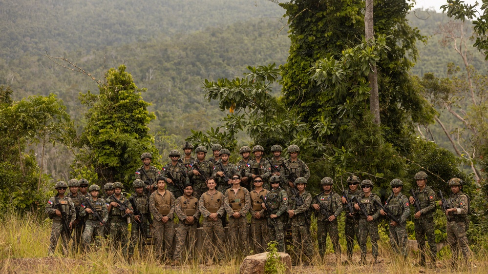 A large group of approximately 25-30 military personnel pose for a group photograph in a tropical jungle/forested setting. The group appears to consist of soldiers from multiple nations, identifiable by different uniform patterns and flag patches, suggesting a joint military exercise or training operation. Several individuals in the front center are wearing lighter-colored uniforms distinct from the camouflage-clad soldiers surrounding them.