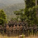 A large group of approximately 25-30 military personnel pose for a group photograph in a tropical jungle/forested setting. The group appears to consist of soldiers from multiple nations, identifiable by different uniform patterns and flag patches, suggesting a joint military exercise or training operation. Several individuals in the front center are wearing lighter-colored uniforms distinct from the camouflage-clad soldiers surrounding them.