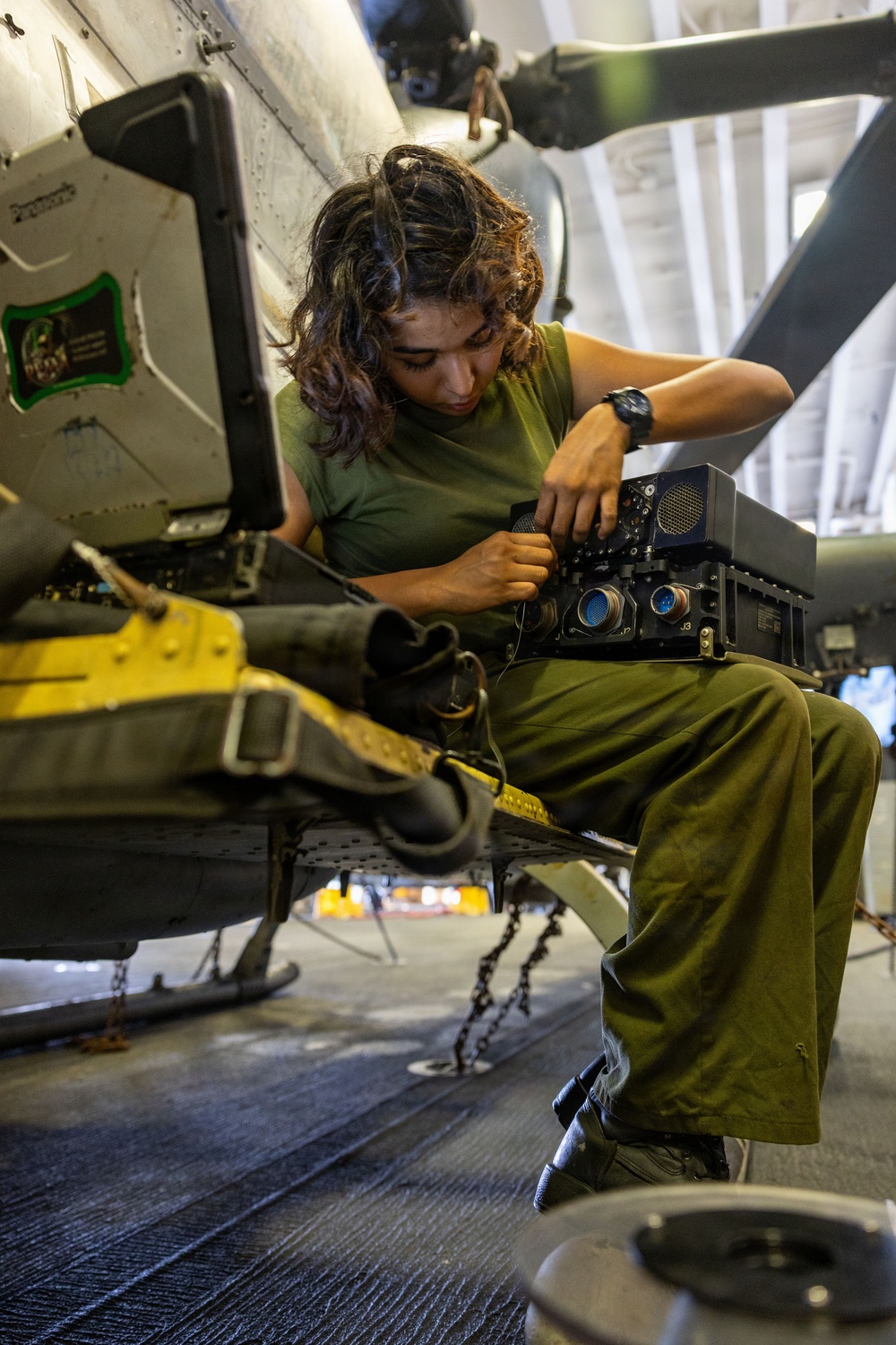 A military technician wearing olive green utilities is performing maintenance or inspection work on avionics or electronic equipment mounted on a helicopter, likely inside a hangar. The individual is seated on the aircraft's step or skid structure, closely examining and handling a black electronic box with visible connector ports. The setting appears to be an indoor aircraft maintenance facility.