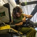 A military technician wearing olive green utilities is performing maintenance or inspection work on avionics or electronic equipment mounted on a helicopter, likely inside a hangar. The individual is seated on the aircraft's step or skid structure, closely examining and handling a black electronic box with visible connector ports. The setting appears to be an indoor aircraft maintenance facility.