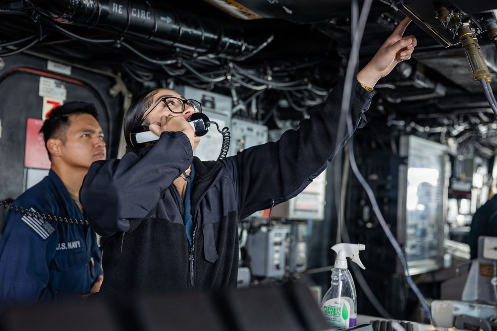 Two U.S. Navy personnel are working inside what appears to be a ship's interior compartment or control space. One sailor is speaking on a corded telephone handset while pointing upward at overhead cables and piping. A second sailor in a U.S. Navy uniform stands nearby observing.