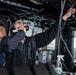 Two U.S. Navy personnel are working inside what appears to be a ship's interior compartment or control space. One sailor is speaking on a corded telephone handset while pointing upward at overhead cables and piping. A second sailor in a U.S. Navy uniform stands nearby observing.