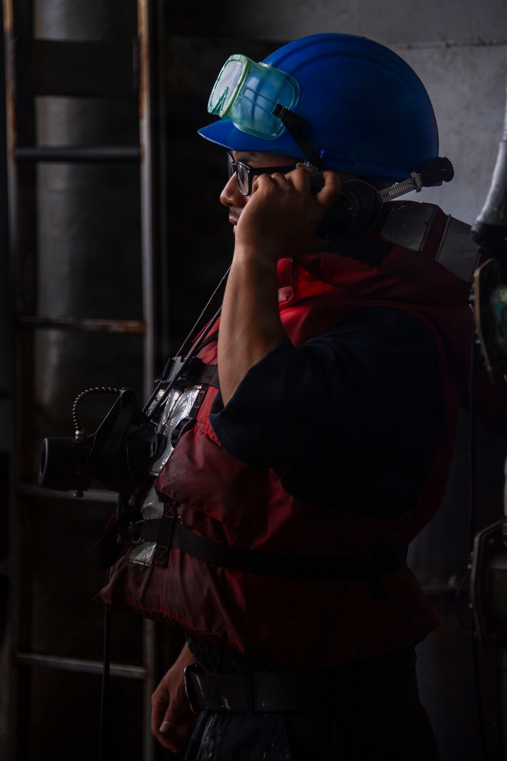 A worker in an industrial environment is wearing a blue hard hat with green safety goggles pushed up on the helmet, along with eyeglasses and a red safety vest or jacket. The individual appears to be holding a communication device (radio or phone handset) to their ear. They are equipped with what appears to be a self-contained breathing apparatus (SCBA) or similar backpack-mounted equipment on their back.