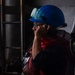A worker in an industrial environment is wearing a blue hard hat with green safety goggles pushed up on the helmet, along with eyeglasses and a red safety vest or jacket. The individual appears to be holding a communication device (radio or phone handset) to their ear. They are equipped with what appears to be a self-contained breathing apparatus (SCBA) or similar backpack-mounted equipment on their back.