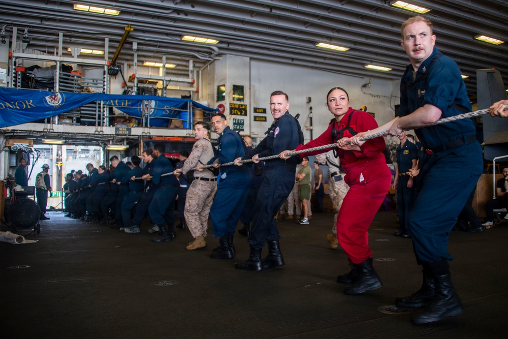 Military and naval personnel are engaged in a tug-of-war competition inside what appears to be the hangar bay of a naval vessel. Participants are wearing a variety of uniforms including Navy coveralls, Marine Corps camouflage, and one person in red coveralls, suggesting a multi-branch or inter-unit event. The setting features industrial overhead lighting, signage, and equipment typical of a ship's interior bay.