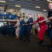 Military and naval personnel are engaged in a tug-of-war competition inside what appears to be the hangar bay of a naval vessel. Participants are wearing a variety of uniforms including Navy coveralls, Marine Corps camouflage, and one person in red coveralls, suggesting a multi-branch or inter-unit event. The setting features industrial overhead lighting, signage, and equipment typical of a ship's interior bay.