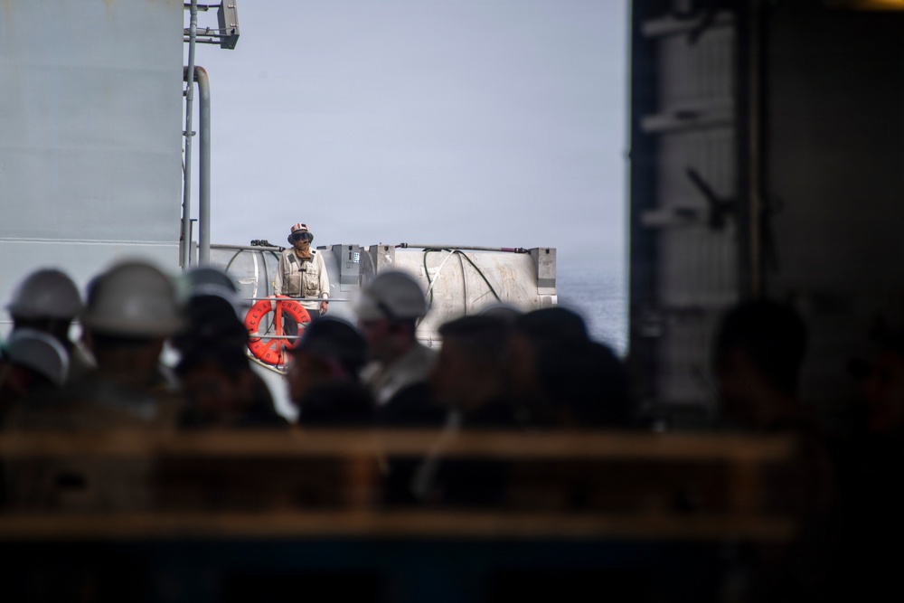 The image shows a group of personnel wearing white hard hats gathered on what appears to be the deck of a large vessel or ship. One individual stands elevated above the group, wearing a helmet and vest, possibly in a supervisory or briefing role. An orange life ring is visible, confirming a maritime setting.