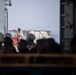 The image shows a group of personnel wearing white hard hats gathered on what appears to be the deck of a large vessel or ship. One individual stands elevated above the group, wearing a helmet and vest, possibly in a supervisory or briefing role. An orange life ring is visible, confirming a maritime setting.