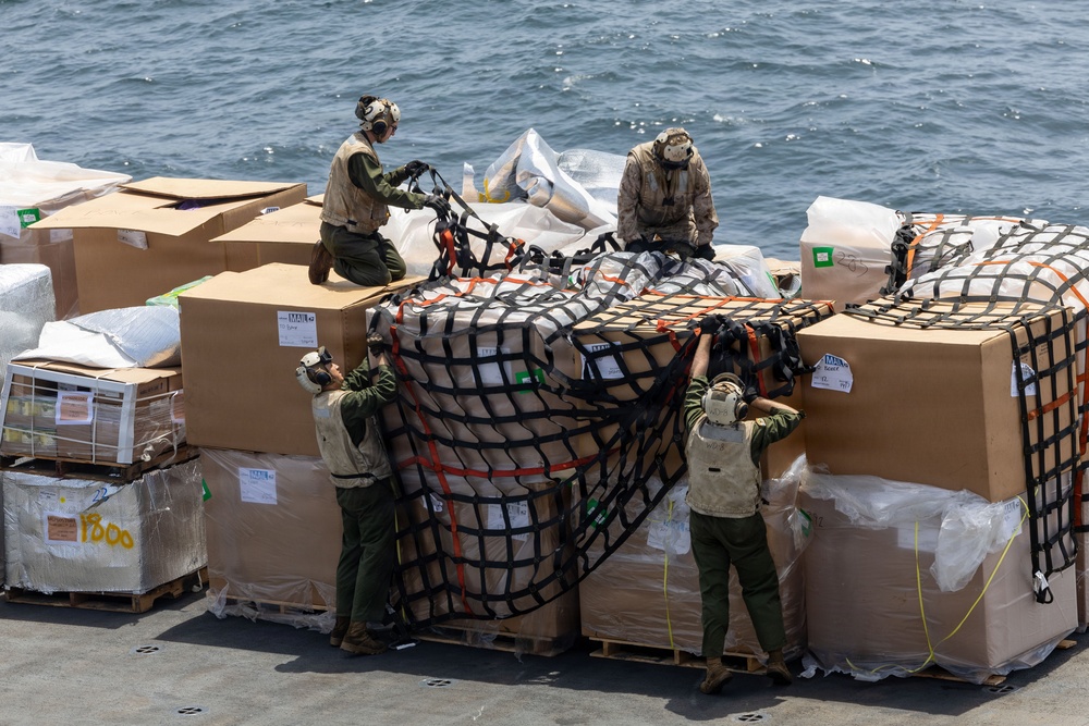Four military personnel in uniform and protective gear are working on a ship's deck to secure or unsecure cargo pallets using cargo nets with orange straps. The pallets are stacked with large cardboard boxes and wrapped goods, situated on what appears to be a naval vessel deck with open ocean visible in the background. The operation appears to be a vertical replenishment or cargo handling evolution.