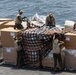 Four military personnel in uniform and protective gear are working on a ship's deck to secure or unsecure cargo pallets using cargo nets with orange straps. The pallets are stacked with large cardboard boxes and wrapped goods, situated on what appears to be a naval vessel deck with open ocean visible in the background. The operation appears to be a vertical replenishment or cargo handling evolution.