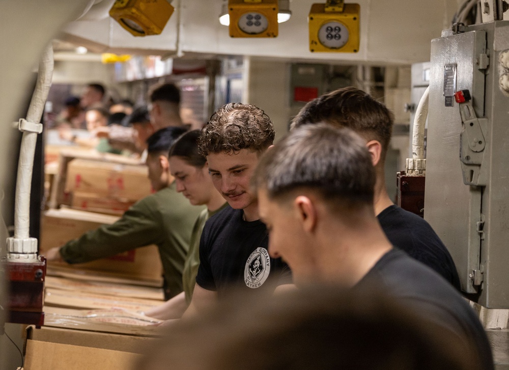 A group of young men are working in a line passing or handling cardboard boxes in what appears to be a confined industrial or military vessel interior. The setting features overhead yellow electrical fixtures and gray metal paneling consistent with a naval ship's interior. The workers are dressed in casual civilian clothing including t-shirts and a green long-sleeve shirt.