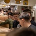 A group of young men are working in a line passing or handling cardboard boxes in what appears to be a confined industrial or military vessel interior. The setting features overhead yellow electrical fixtures and gray metal paneling consistent with a naval ship's interior. The workers are dressed in casual civilian clothing including t-shirts and a green long-sleeve shirt.