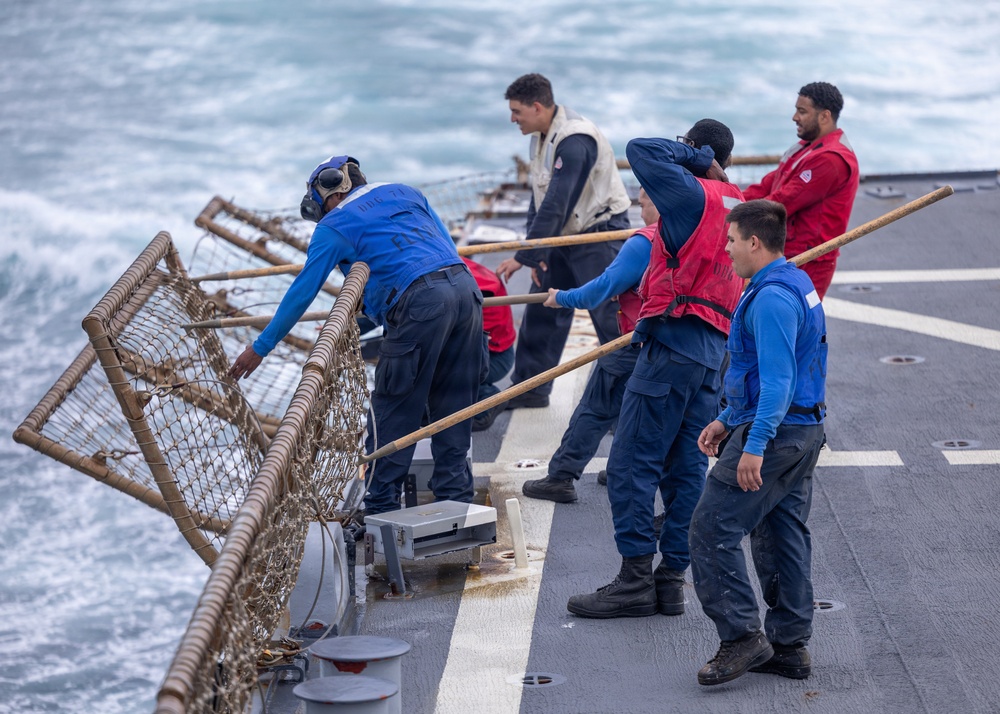 A group of naval personnel are working on the deck of a military vessel at sea, handling what appears to be a cargo net or safety netting along the ship's railing. The crew members are wearing a mix of blue coveralls and red safety vests, with some holding wooden-handled poles or boat hooks. The sea conditions appear rough, with visible white-capped waves alongside the ship.