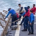 A group of naval personnel are working on the deck of a military vessel at sea, handling what appears to be a cargo net or safety netting along the ship's railing. The crew members are wearing a mix of blue coveralls and red safety vests, with some holding wooden-handled poles or boat hooks. The sea conditions appear rough, with visible white-capped waves alongside the ship.