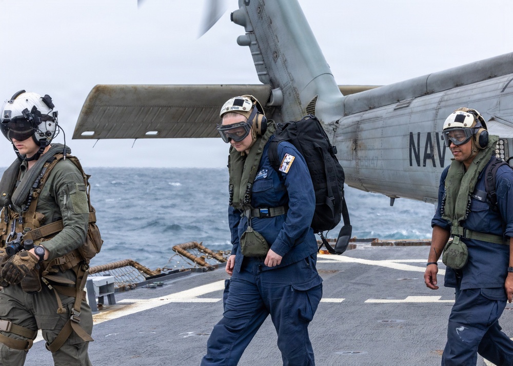 Three military personnel are walking across a naval vessel's flight deck with a Navy helicopter visible in the background. The individuals are wearing different uniforms and protective gear, suggesting a multi-service or mixed-role operation. The scene takes place at sea under overcast conditions.
