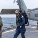 Three military personnel are walking across a naval vessel's flight deck with a Navy helicopter visible in the background. The individuals are wearing different uniforms and protective gear, suggesting a multi-service or mixed-role operation. The scene takes place at sea under overcast conditions.