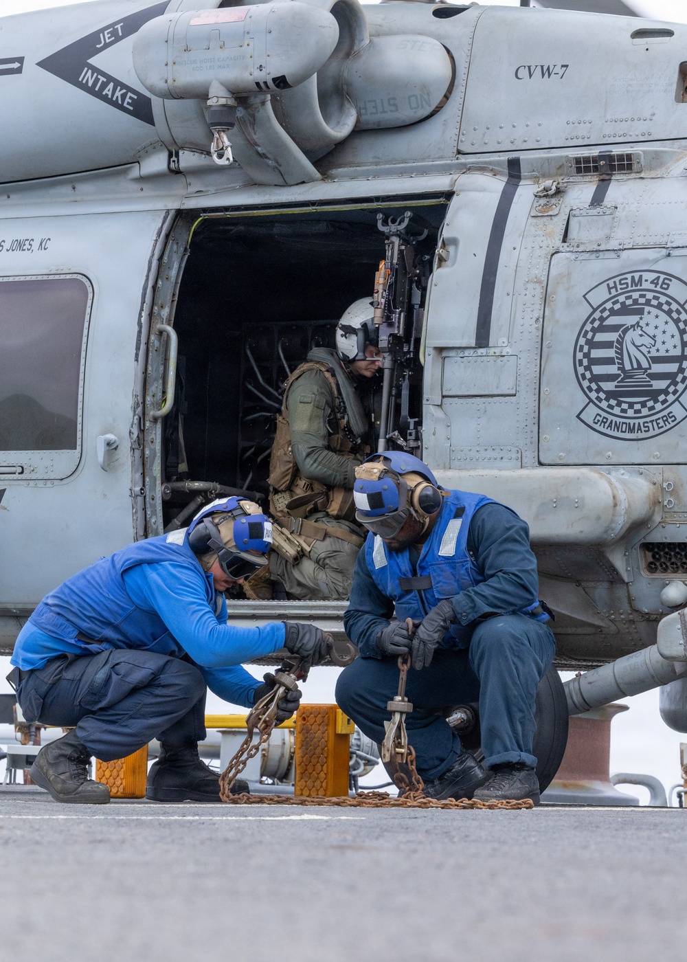 Two naval deck crew members in blue jerseys and cranial helmets are securing tie-down chains to a helicopter's landing gear on what appears to be an aircraft carrier flight deck. The helicopter is marked with 'HSM-46 Grandmasters' and 'CVW-7' insignia, indicating it belongs to Helicopter Maritime Strike Squadron 46. A third person in tactical gear and a flight helmet is seated inside the open cabin door of the helicopter.