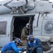 Two naval deck crew members in blue jerseys and cranial helmets are securing tie-down chains to a helicopter's landing gear on what appears to be an aircraft carrier flight deck. The helicopter is marked with 'HSM-46 Grandmasters' and 'CVW-7' insignia, indicating it belongs to Helicopter Maritime Strike Squadron 46. A third person in tactical gear and a flight helmet is seated inside the open cabin door of the helicopter.