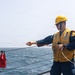 A naval or maritime worker wearing a yellow hard hat and safety vest is handling lines/ropes on the deck of a ship at sea. The individual appears to be working with a red line and a connector or shackle, likely involved in a replenishment-at-sea or line-handling operation. A red bag and yellow line are visible attached to a railing post in the foreground.