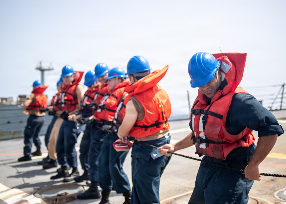 A group of naval personnel are lined up on the deck of a ship, pulling a rope in a coordinated line formation. All individuals are wearing orange life vests and blue hard hats, indicating a safety-compliant maritime operation. The activity appears to be a line-handling or mooring drill conducted at sea.