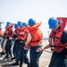 A group of naval personnel are lined up on the deck of a ship, pulling a rope in a coordinated line formation. All individuals are wearing orange life vests and blue hard hats, indicating a safety-compliant maritime operation. The activity appears to be a line-handling or mooring drill conducted at sea.