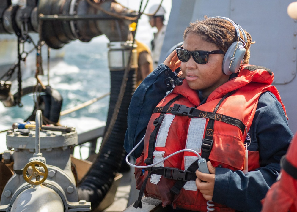A naval or coast guard personnel wearing an orange life vest and gray communication headset is standing on the deck of a vessel at sea. The individual is holding a communication device and appears to be monitoring or coordinating an operation. Maritime equipment including hoses, valves, and rigging hardware are visible in the foreground.