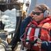 A naval or coast guard personnel wearing an orange life vest and gray communication headset is standing on the deck of a vessel at sea. The individual is holding a communication device and appears to be monitoring or coordinating an operation. Maritime equipment including hoses, valves, and rigging hardware are visible in the foreground.