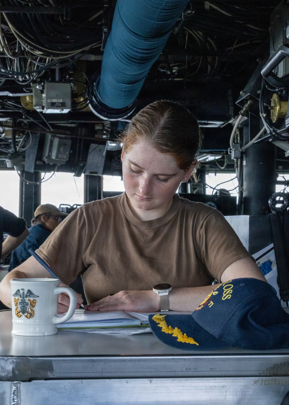 A young female sailor in a tan/coyote brown U.S. Navy t-shirt is writing or reviewing documents at a workstation aboard a naval vessel. She is seated on what appears to be the bridge or a command area, surrounded by cables, ducts, and shipboard equipment. A U.S. Navy coffee mug and a navy blue officer's cover (cap) with gold insignia are visible on the surface beside her.