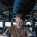 A young female sailor in a tan/coyote brown U.S. Navy t-shirt is writing or reviewing documents at a workstation aboard a naval vessel. She is seated on what appears to be the bridge or a command area, surrounded by cables, ducts, and shipboard equipment. A U.S. Navy coffee mug and a navy blue officer's cover (cap) with gold insignia are visible on the surface beside her.