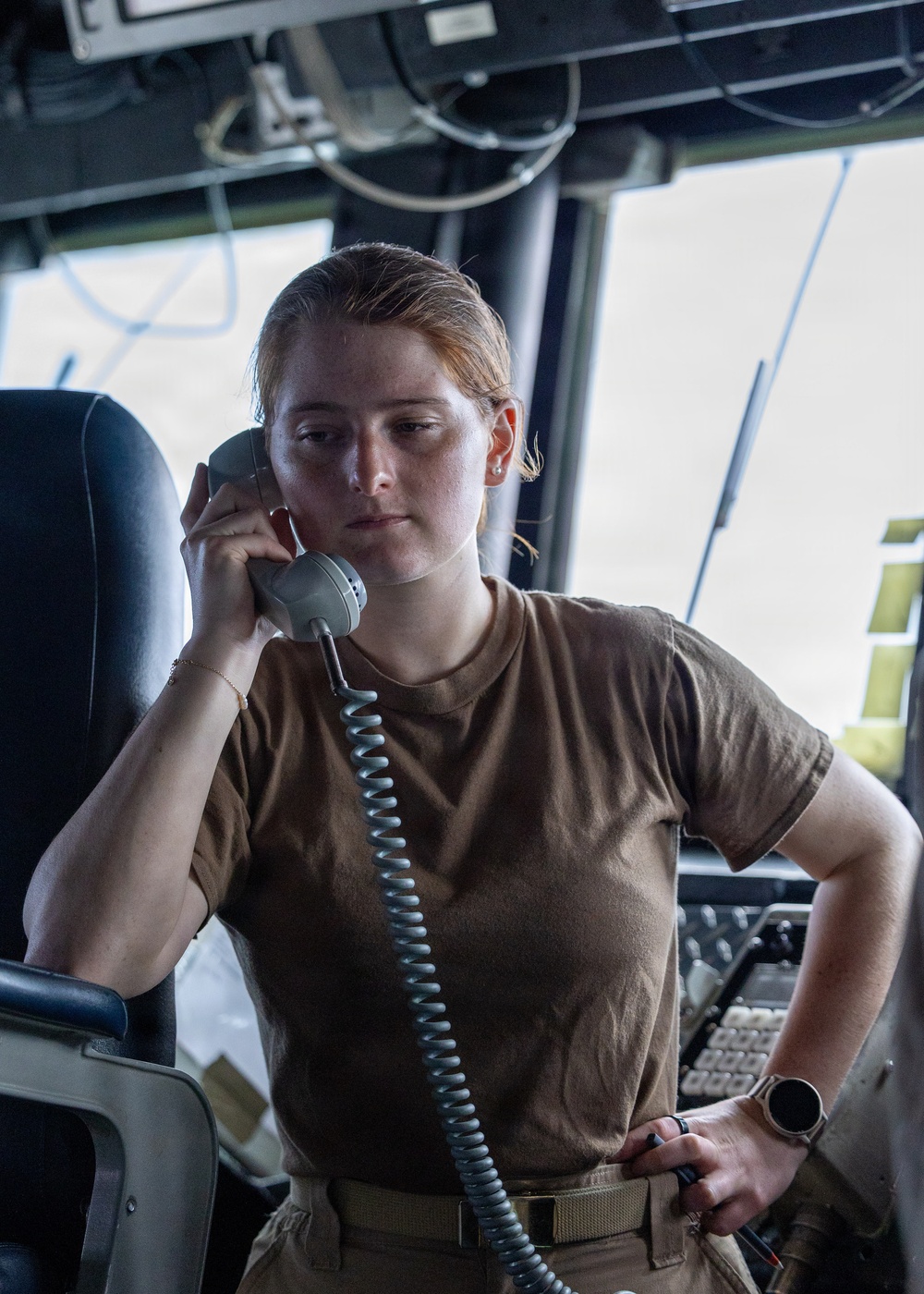 A young woman in military-style attire is using a corded telephone handset in what appears to be a ship's bridge or command center. She is wearing a tan/coyote brown t-shirt with a matching belt, and appears focused and attentive. The setting is filled with communications and navigation equipment consistent with a naval vessel.