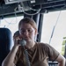 A young woman in military-style attire is using a corded telephone handset in what appears to be a ship's bridge or command center. She is wearing a tan/coyote brown t-shirt with a matching belt, and appears focused and attentive. The setting is filled with communications and navigation equipment consistent with a naval vessel.