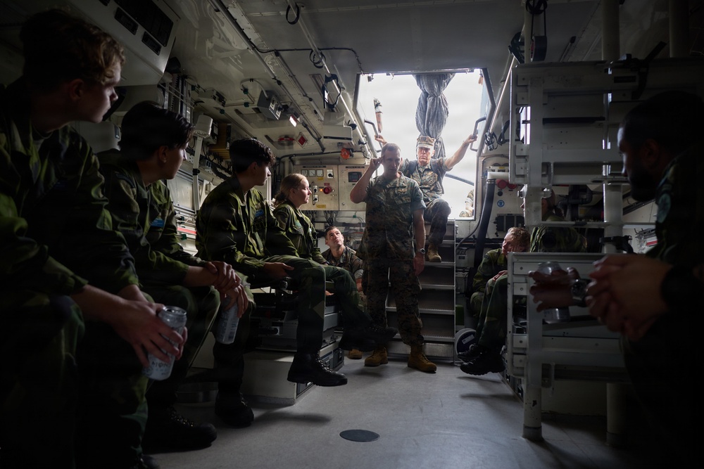 Military personnel are seated inside what appears to be the interior of a military vehicle or vessel, likely an amphibious assault vehicle or similar transport. A standing service member in US Marine Corps-style uniform appears to be briefing or instructing the group, while others seated on both sides listen attentively. The personnel are wearing a mix of camouflage uniforms suggesting a multinational or joint military exercise.