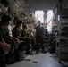 Military personnel are seated inside what appears to be the interior of a military vehicle or vessel, likely an amphibious assault vehicle or similar transport. A standing service member in US Marine Corps-style uniform appears to be briefing or instructing the group, while others seated on both sides listen attentively. The personnel are wearing a mix of camouflage uniforms suggesting a multinational or joint military exercise.