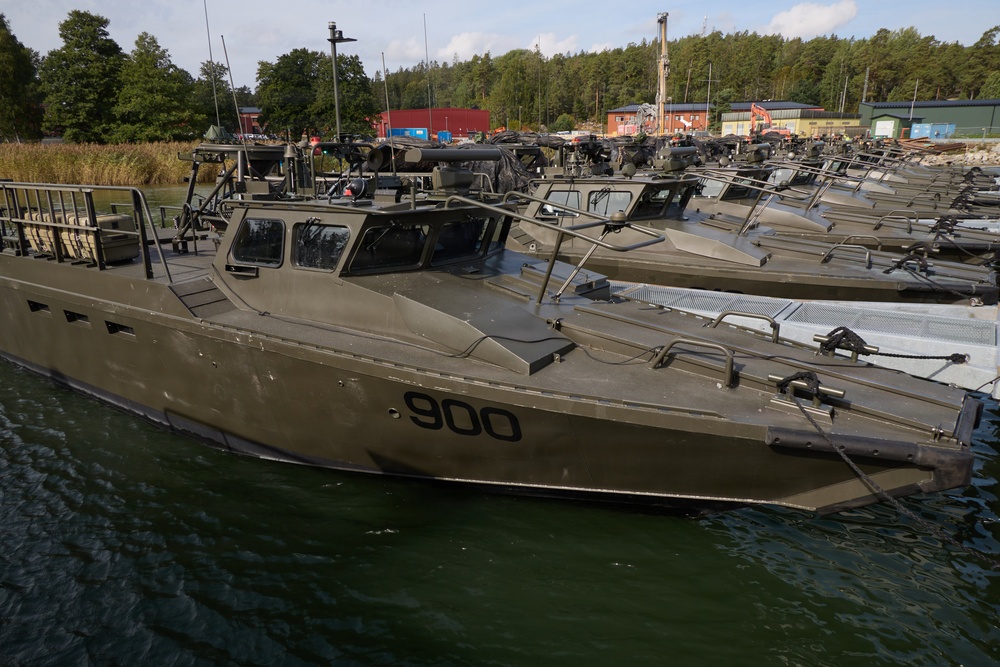 A fleet of military patrol/assault boats painted in olive drab/military green are moored at a dock. The nearest vessel is clearly marked with the hull number '900'. The setting appears to be a military naval base surrounded by forested terrain, with industrial and storage buildings visible in the background.