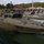A fleet of military patrol/assault boats painted in olive drab/military green are moored at a dock. The nearest vessel is clearly marked with the hull number '900'. The setting appears to be a military naval base surrounded by forested terrain, with industrial and storage buildings visible in the background.