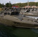 A fleet of military patrol/assault boats painted in olive drab/military green are moored at a dock. The nearest vessel is clearly marked with the hull number '900'. The setting appears to be a military naval base surrounded by forested terrain, with industrial and storage buildings visible in the background.